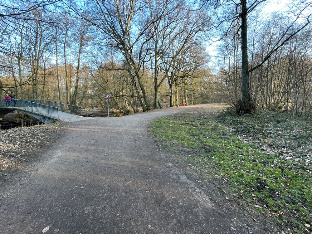 Weg durch einen Park, links Fußgängerbrücke über ein Gewässer