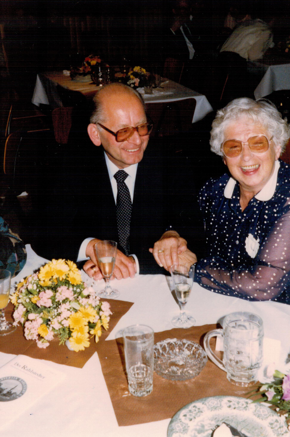 Älterer Mann und Frau mit weißen Haaren sitzen an einem Tisch und die Frau lacht in die Kamera